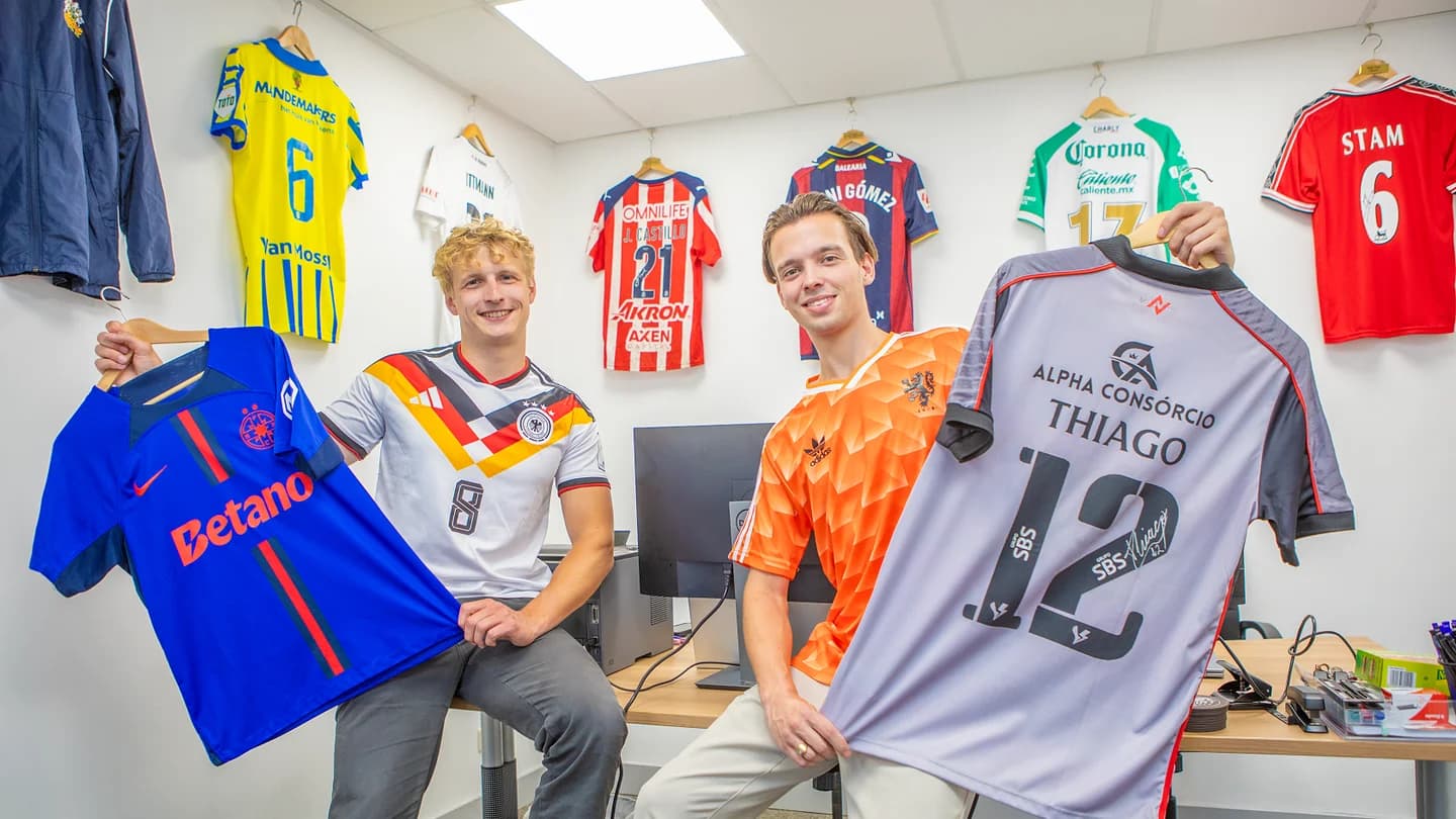 The two Shirt Circle founders in their office, each holding a football shirt, with more shirts on the wall behind them